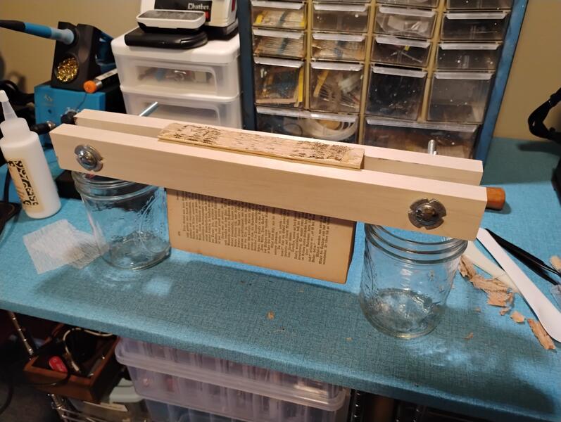 The photo shows a simple text block clamp I made, the text block of the book secured in the clamp while fresh archival glue dries to reinforce the spine and apply new mull. The clamp is propped up on two wide-mouth mason jars because they were convenient.