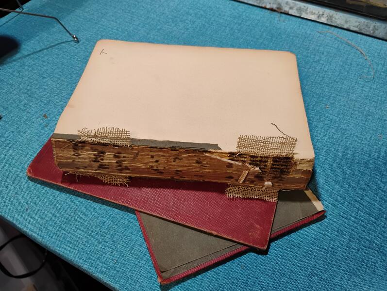 A photo of the book being worked on in this project. The spine is completely exposed, and the remaining cover panels have been carefully removed the rest of the way, althoug they were hanging on by mere threads as it was. The text block sits atop the removed cover panels, ready to be re-cased.